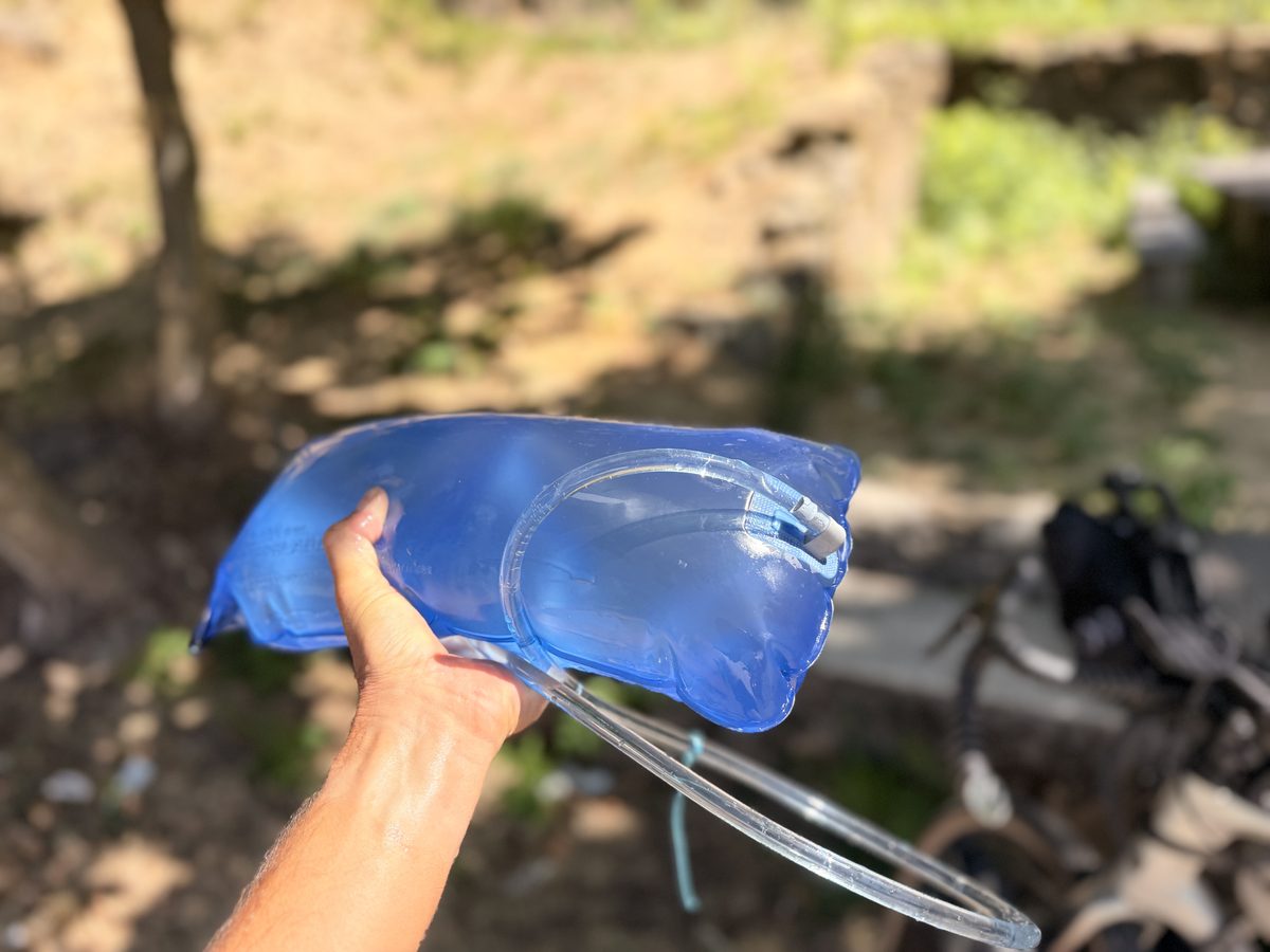 Hand hält eine gefüllte 3-Liter-Trinkblase mit Trinkschlauch, im Hintergrund unscharf das Fahrrad