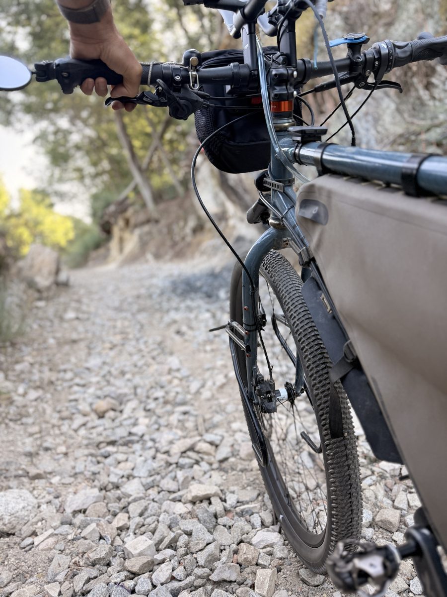 Blick über den schmalen MTB-Flatbar auf eine Schotterpiste, Hand am Griff
